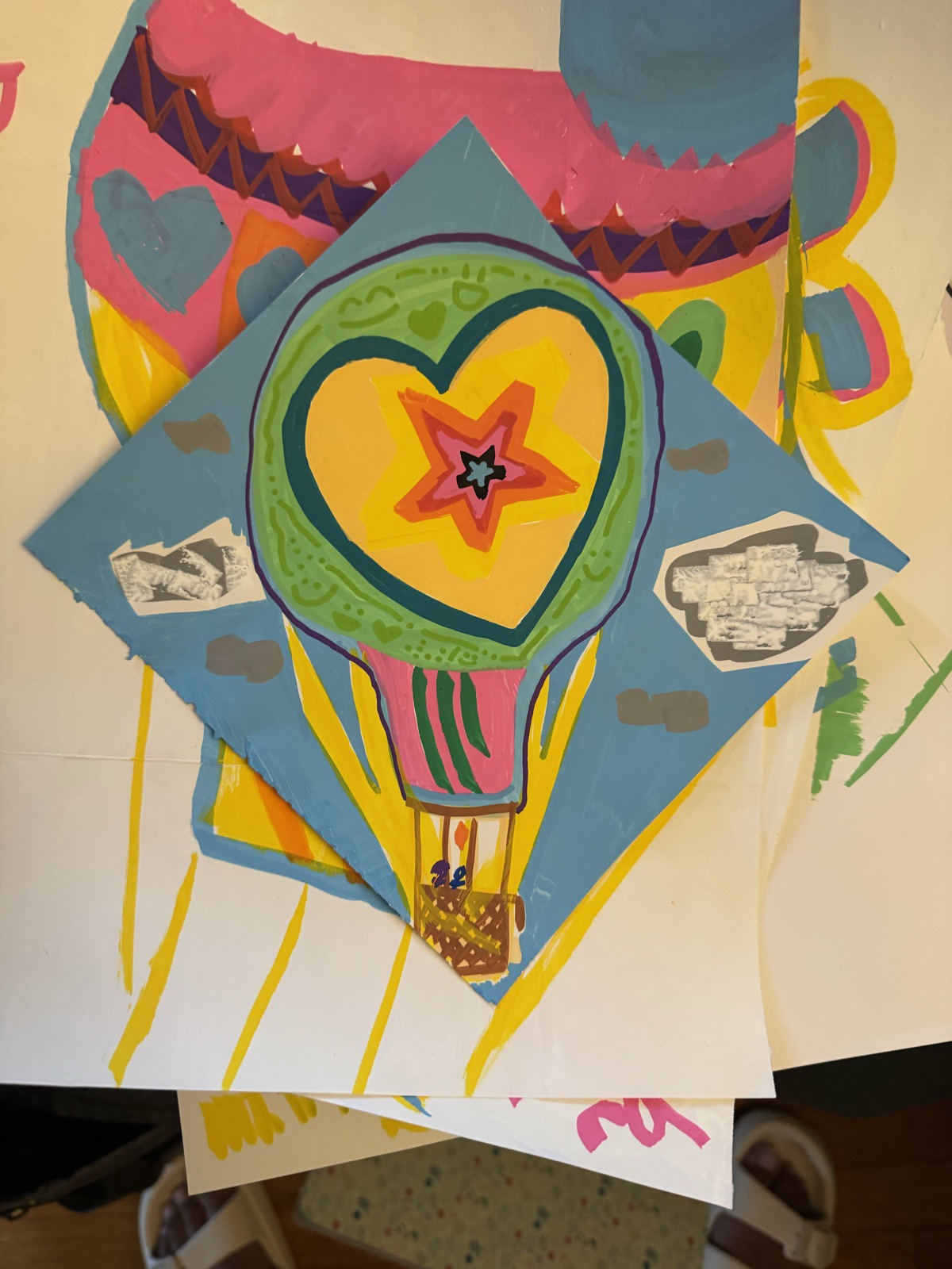 Hot-air balloon collage seen straight on: a green-ringed balloon with a heart and a star inside, on a blue diamond of paper with grayscale cutouts, a woven basket at the bottom