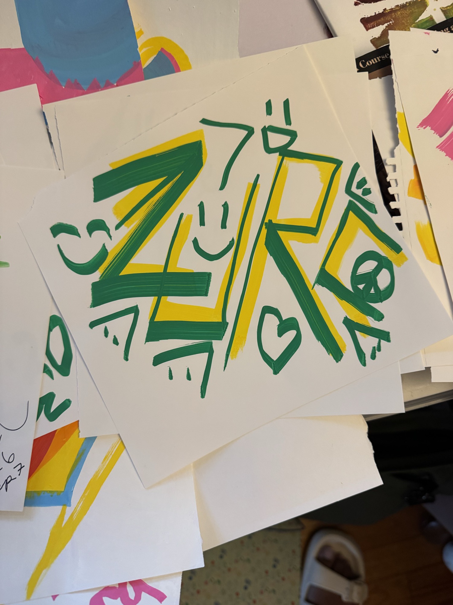 ZURC hand-lettered in green marker over yellow, with a peace sign, hearts, and small faces, on white paper laid over painted sheets